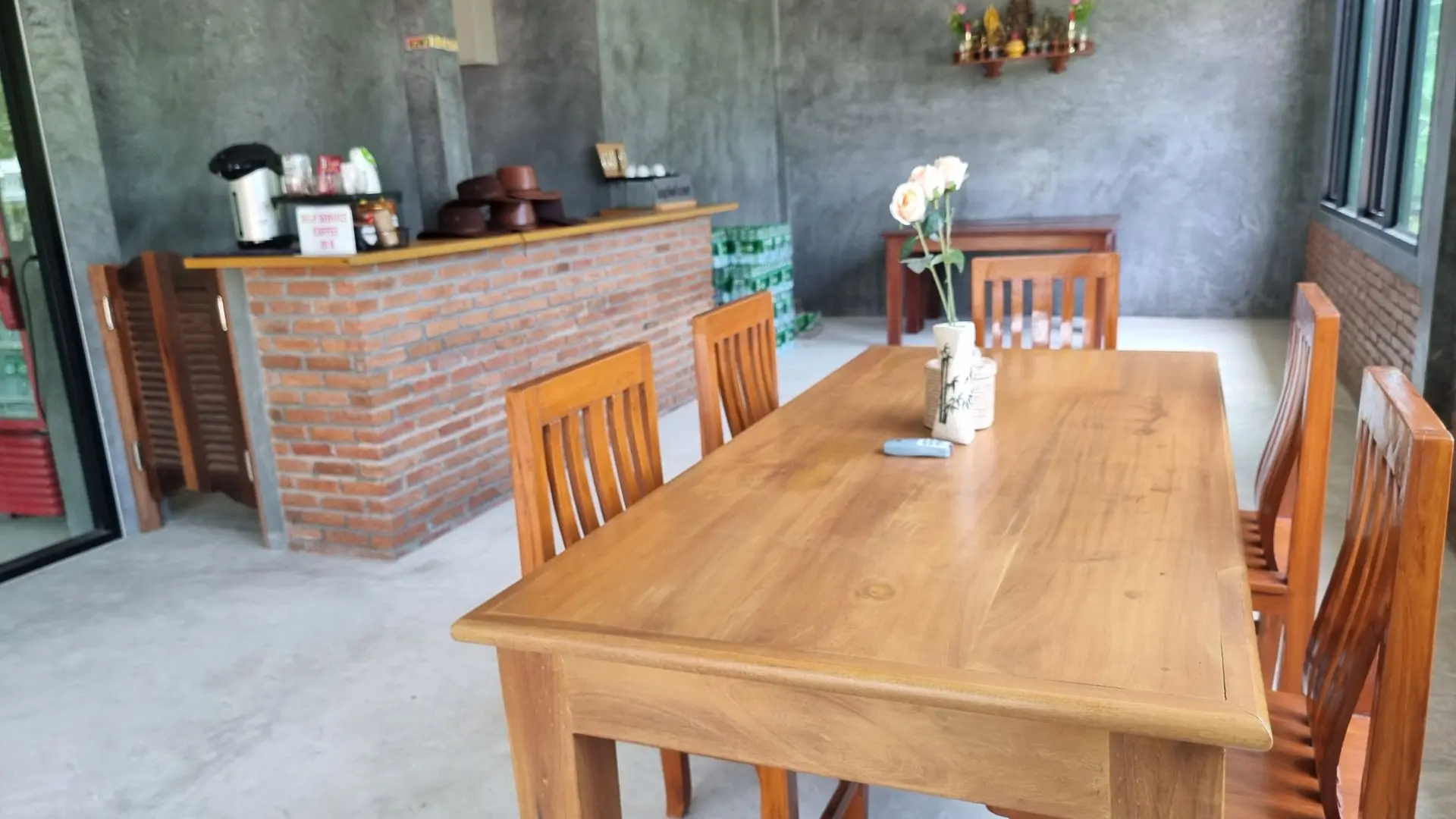 Mesa e cadeiras de madeira numa sala de cimento com balcão de tijolo, café em autosserviço e chapéus empilhados