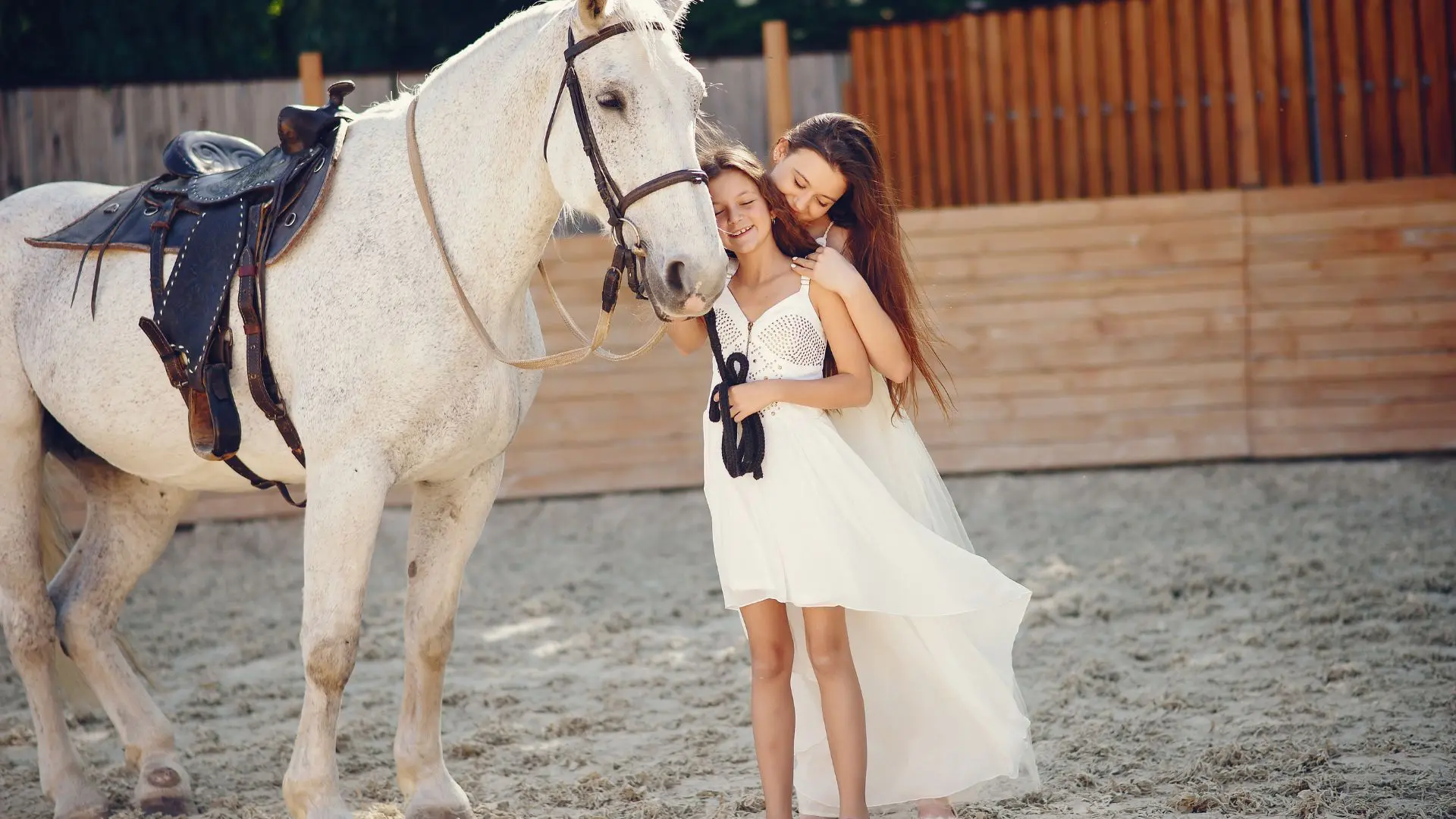 Duas raparigas de vestido branco junto a um cavalo branco selado num picadeiro de areia