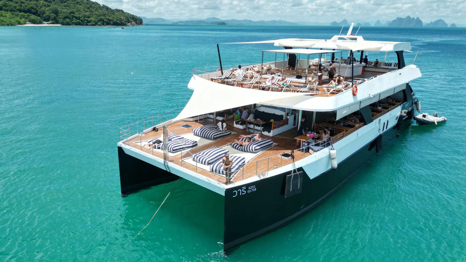 Catamarã preto e branco fundeado em água turquesa, com gente a apanhar sol no convés e ilhéus calcários ao fundo