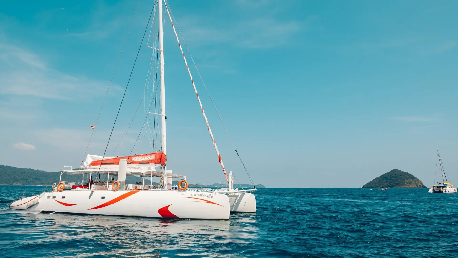 White catamaran with red stripes sailing on the open sea, with a green islet in the background