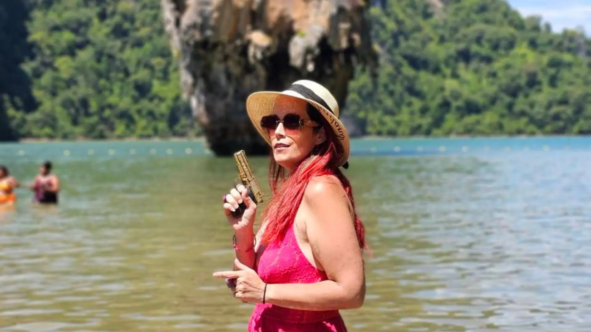 Mulher de fúcsia, cabelo vermelho, óculos de sol e chapéu de palha posa com uma pistola dourada de brincar ante Khao Ta-Pu