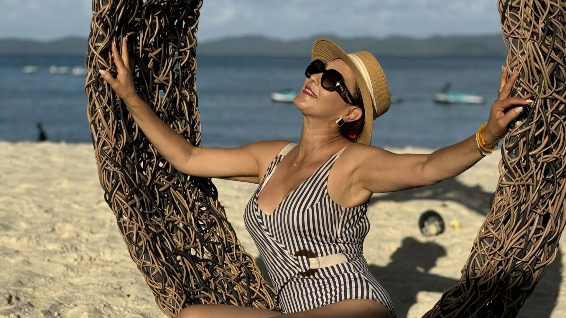 De óculos de sol e chapéu, uma mulher recosta-se num cadeirão entrançado com o mar azul atrás