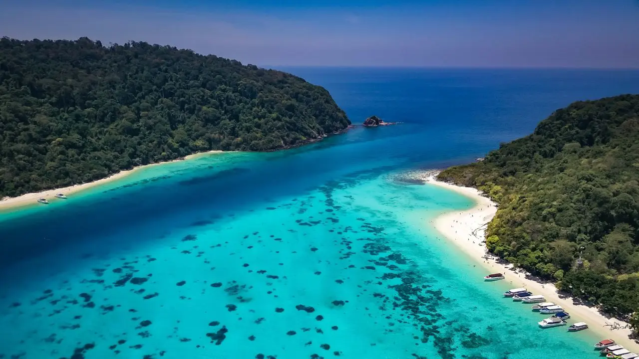Vista aérea do canal pouco fundo entre duas ilhas arborizadas, com corais escuros, praias brancas e lanchas