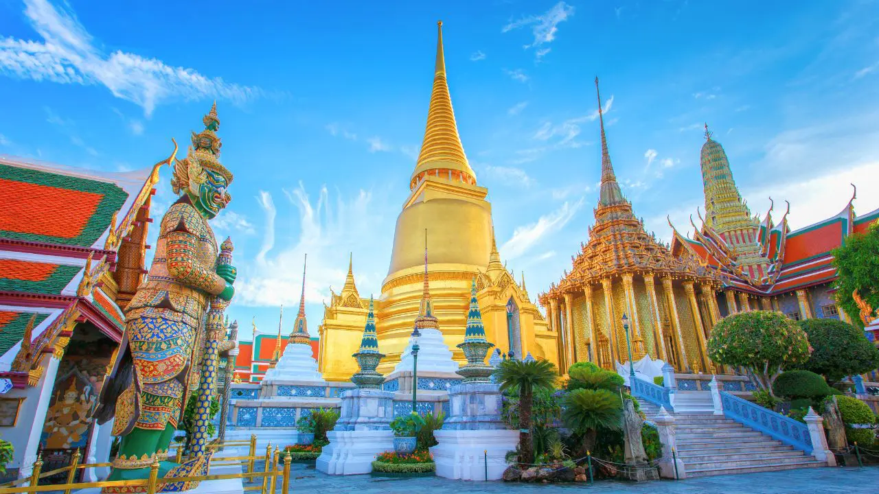 Chedi dourado do Wat Phra Kaew junto a templos de telhados vermelhos e um yaksha verde de guarda sob o céu azul
