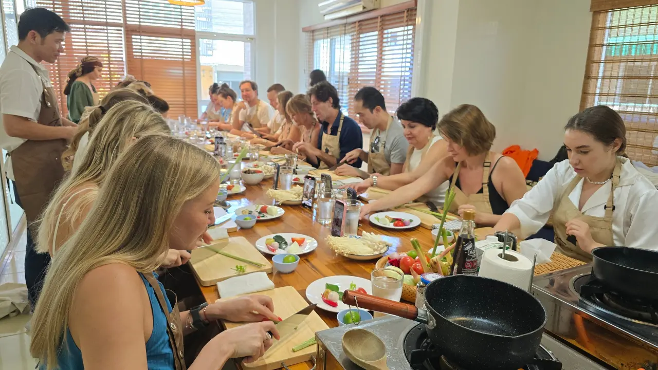 Cooking Class Thai Food in Phuket 6 Large group of students in aprons preparing ingredients at a long wooden table, with an instructor standing