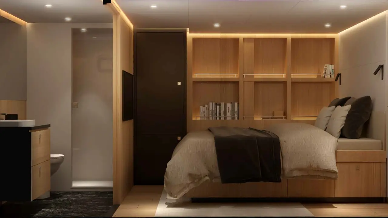 Cabina con cama de edredón beis y manta marrón, estantería con libros, armario oscuro y baño con ducha al lado