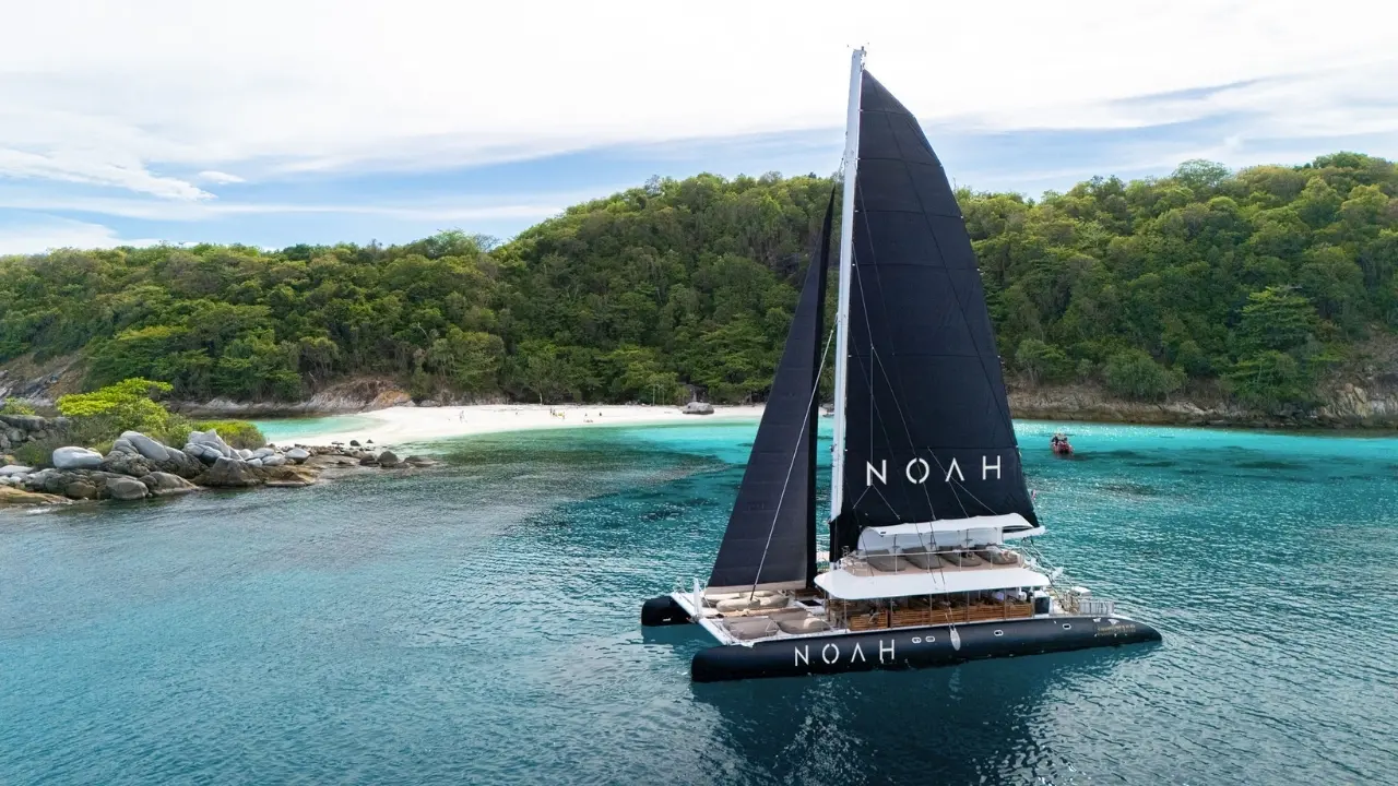 Catamarã de velas pretas em águas turquesa frente a uma enseada de areia branca rodeada de colinas com selva