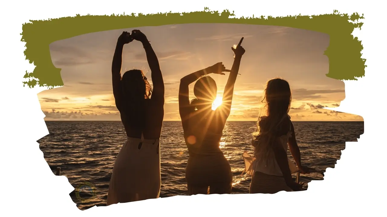Silhuetas de três amigas ao entardecer: uma junta as mãos no ar, outra aponta para o céu com uma taça e outra olha o mar