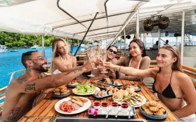 Cinco amigos brindan con espumoso ante una mesa llena de sushi, ensalada y tartaletas, con barcos fondeados detrás